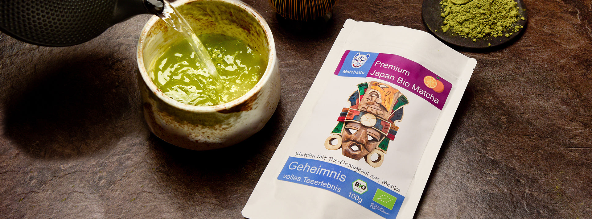 Matchatto Bio-Matcha mit Orangenöl aus Mexiko – Matchapulver, Teeschale mit aufgegossenem Matcha und Verpackung mit bunter Maske.