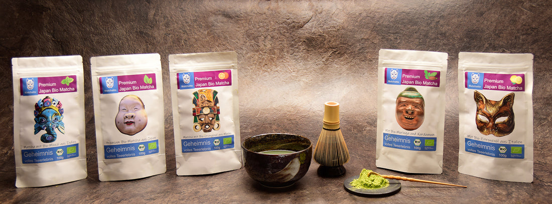 Matchatto Bio-Matcha Sorten in 100g-Packung mit Tama-Masken – kunstvoll inszeniert mit Matchaschale, Chasen und Matchapulver auf Naturstein.