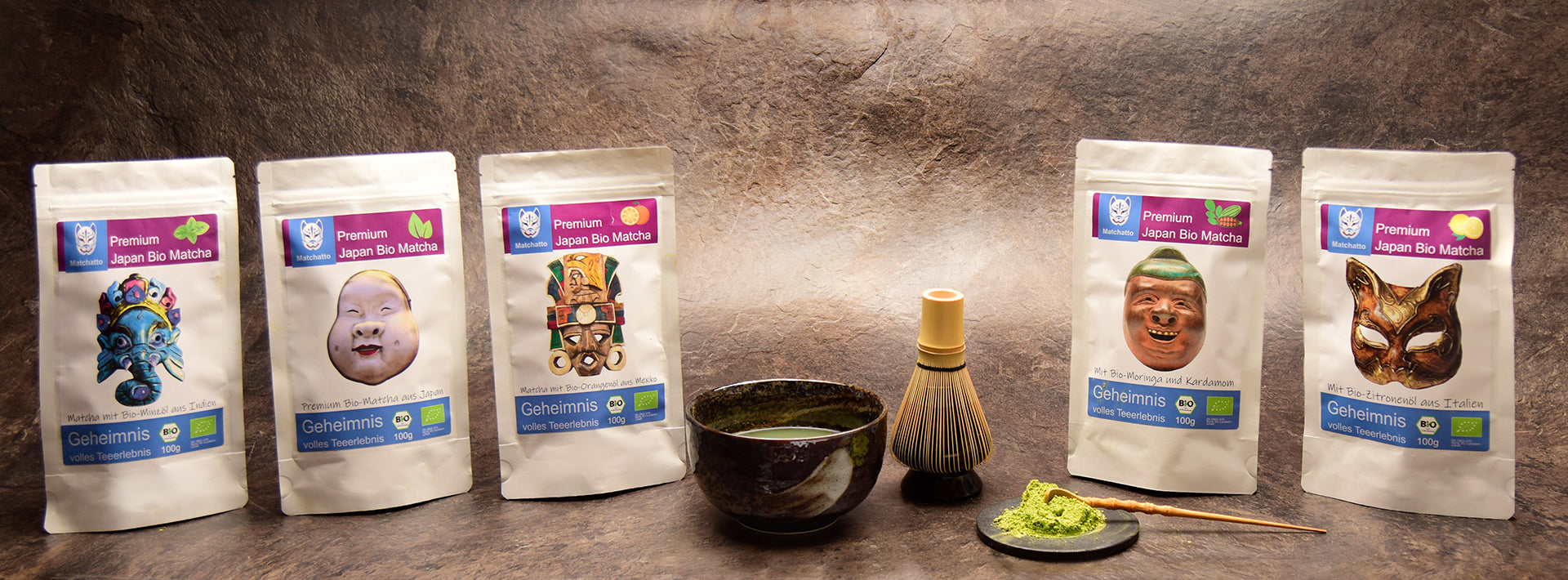 Matchatto Bio-Matcha Sorten in 100g-Packung mit Tama-Masken – kunstvoll inszeniert mit Matchaschale, Chasen und Matchapulver auf Naturstein.