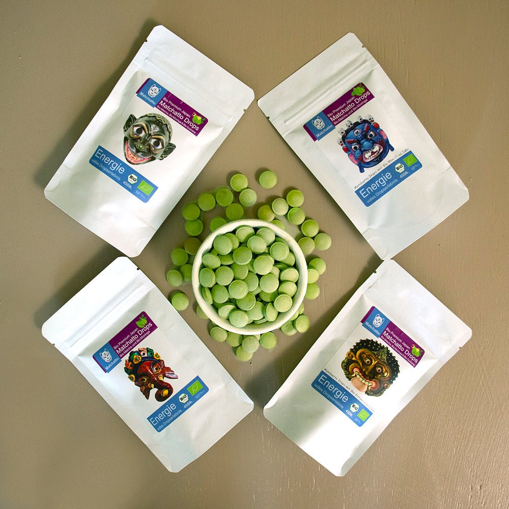 Matchatto Bio Matcha Drops Bundle mit 4 Sorten in weißen Beuteln, rund arrangiert um eine Schale mit grünen Presslingen, Energie-Booster mit Moringa und Minze.