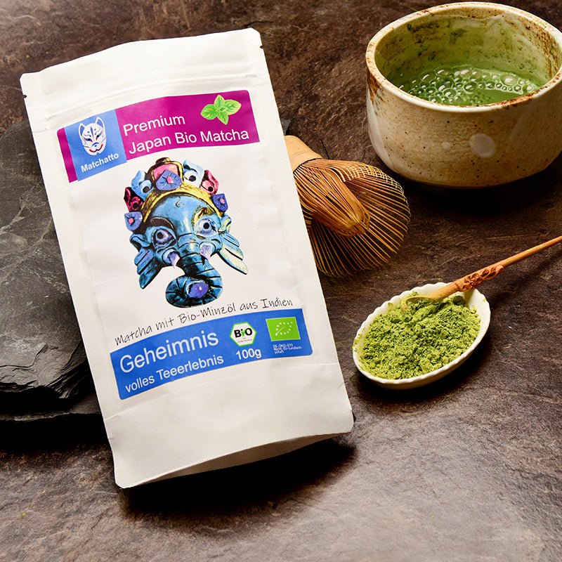 Matchatto Bio Matcha Tee mit Bio-Minze aus Indien in 100g-Beutel, verziert mit blauer Elefantenmaske, daneben Matchaschale, Bambusbesen und Löffel mit grünem Pulver auf dunklem Naturstein.