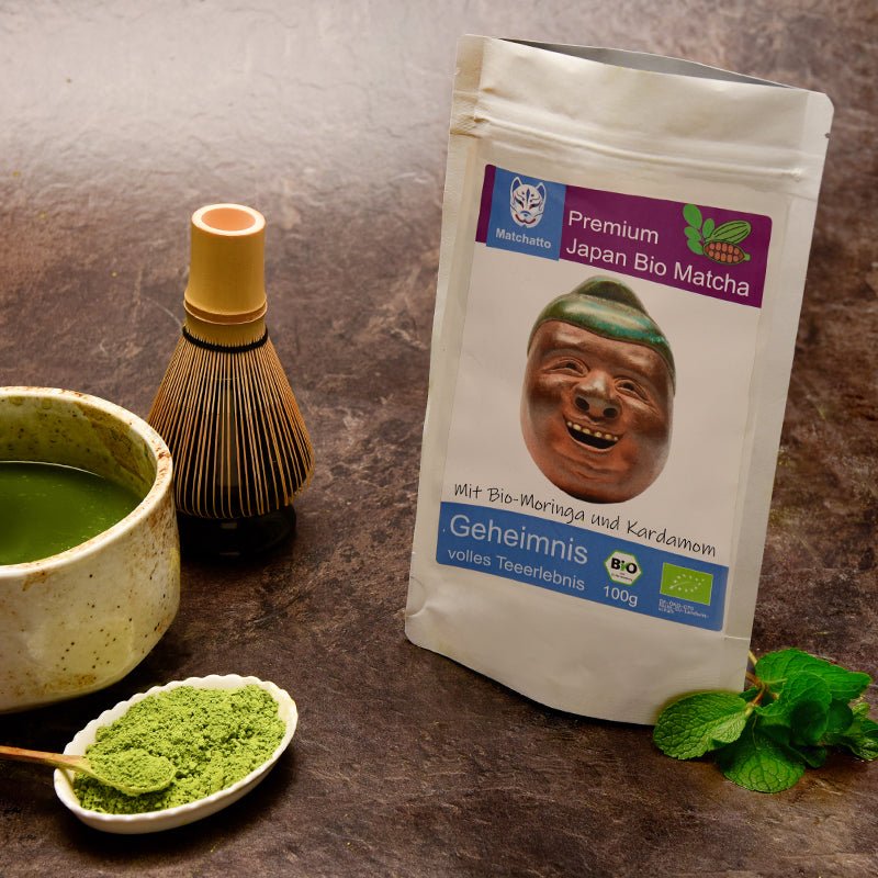 Matchatto Bio-Matcha mit Moringa und Kardamom: Stehender Produktbeutel vor einer Matcha-Schale, Bambusbesen und einer Portion grünem Pulver auf einem Löffel. Daneben frische Minzblätter – eine harmonische Szene für natürliche Balance und Genuss.
