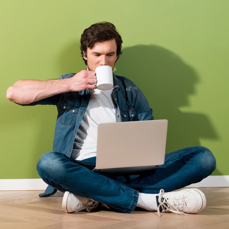 Ein Mann sitzt entspannt angelehnt an eine grüne Wand, mit Laptop auf dem Schoß und trinkt aus einer Tasse.