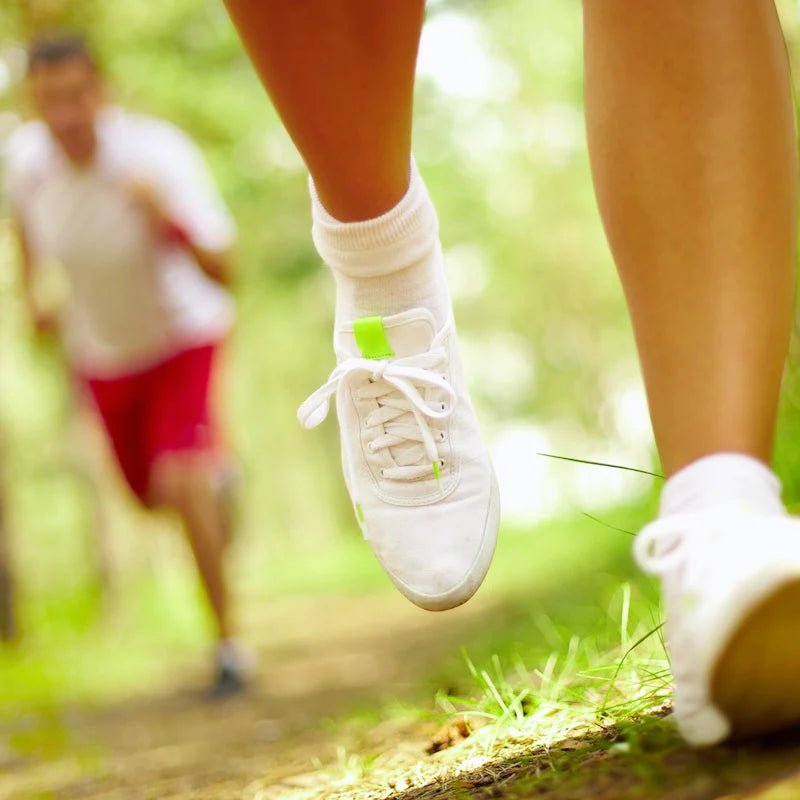 Nahaufnahme von weißen Sportschuhen beim Laufen auf einem Waldweg – im Hintergrund eine weitere Person in Bewegung.