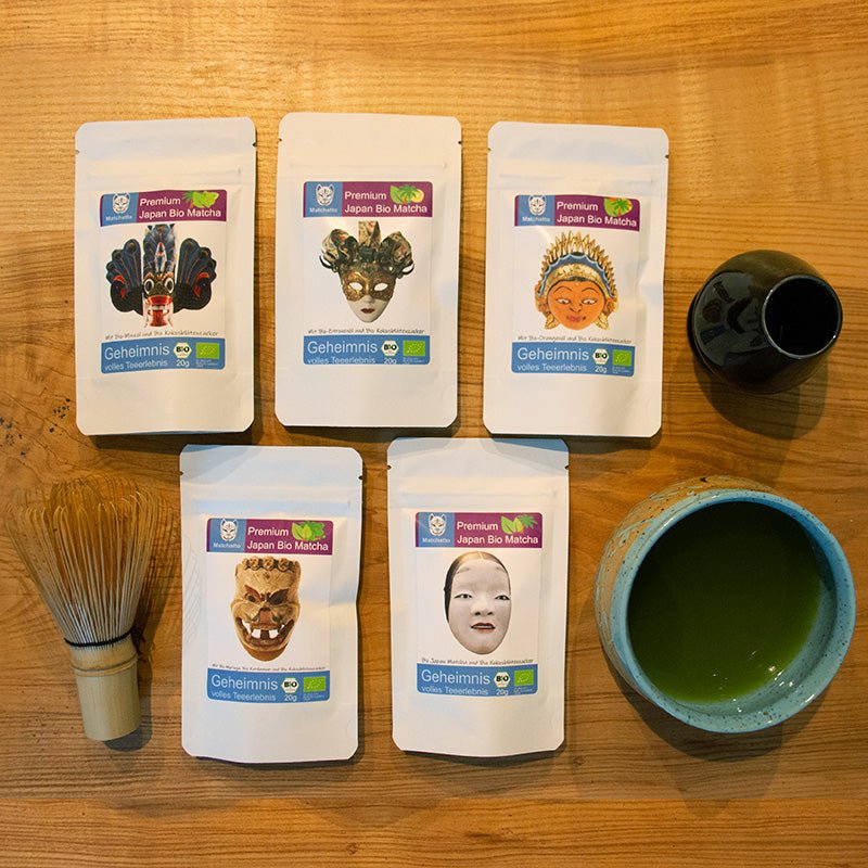 Matchatto Probierpaket mit 6 Sorten Bio Matcha Tee in weißen Beuteln, verziert mit kunstvollen Maskenmotiven, arrangiert auf einem Holztisch mit Matcha-Schale und Bambusbesen.