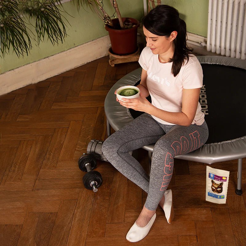 Frau in Sportkleidung sitzt nach dem Training mit Matcha-Tee auf Trampolin – neben ihr Hanteln und Matchatto Bio-Matcha mit Zitronenöl.