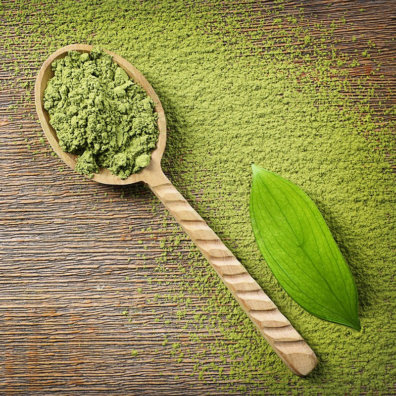 Feines Bio Matcha Tee Pulver in Holzlöffel auf rustikalem Holzhintergrund mit grünem Blatt – naturbelassene Superfood-Zutat aus Japan.