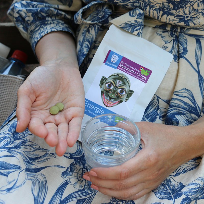 Frau hält Matchatto Bio Matcha Drops und ein Glas Wasser in der Hand – praktische Einnahme unterwegs, Presslingsbeutel mit Bio Matcha und Bio Moringa und Bio Guarana im Hintergrund sichtbar.