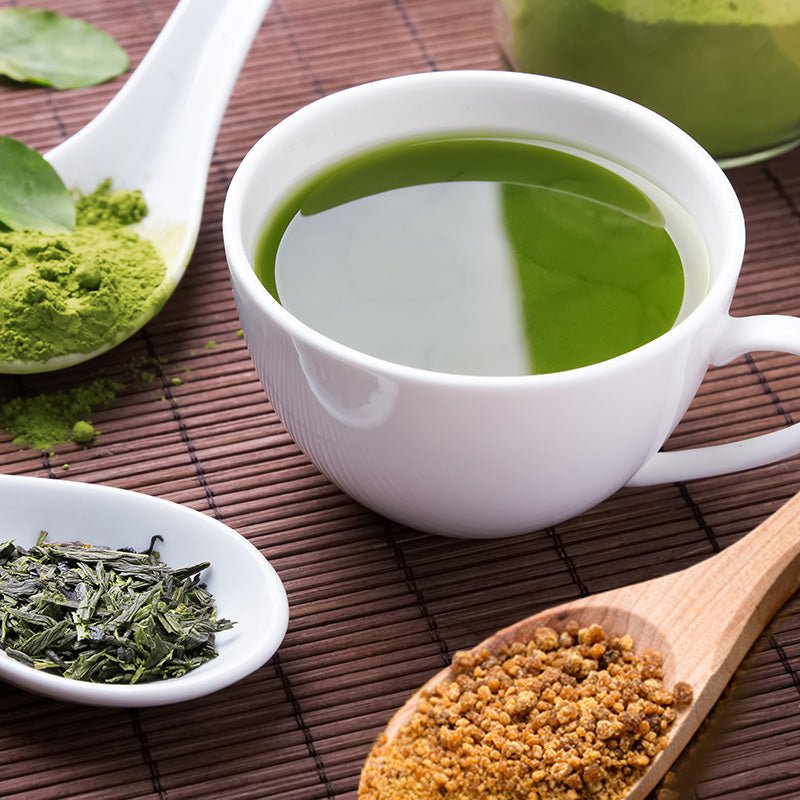 Tasse mit leuchtend grünem Matcha-Tee auf Bambusmatte, umgeben von Matcha-Pulver, grünen Teeblättern und einem Löffel mit Kokosblütenzucker – natürliche Zutaten für bewussten Teegenuss.