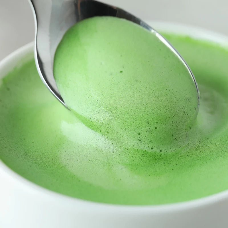 Nahaufnahme einer Tasse mit cremigem Matcha Latte, grünem Schaum und Löffel – vollmundiges Teeerlebnis mit Bio-Matcha.